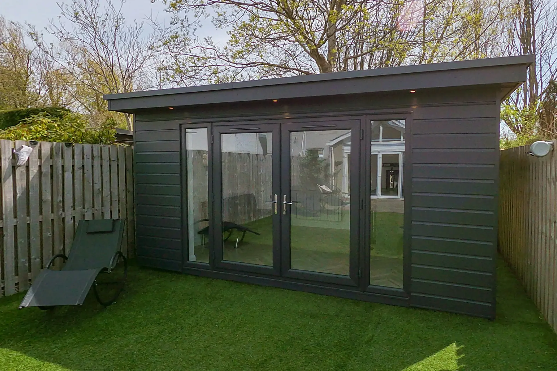 Dark grey wooden garden shed with large glass doors, fenced yard and a lounge chair on the lawn.